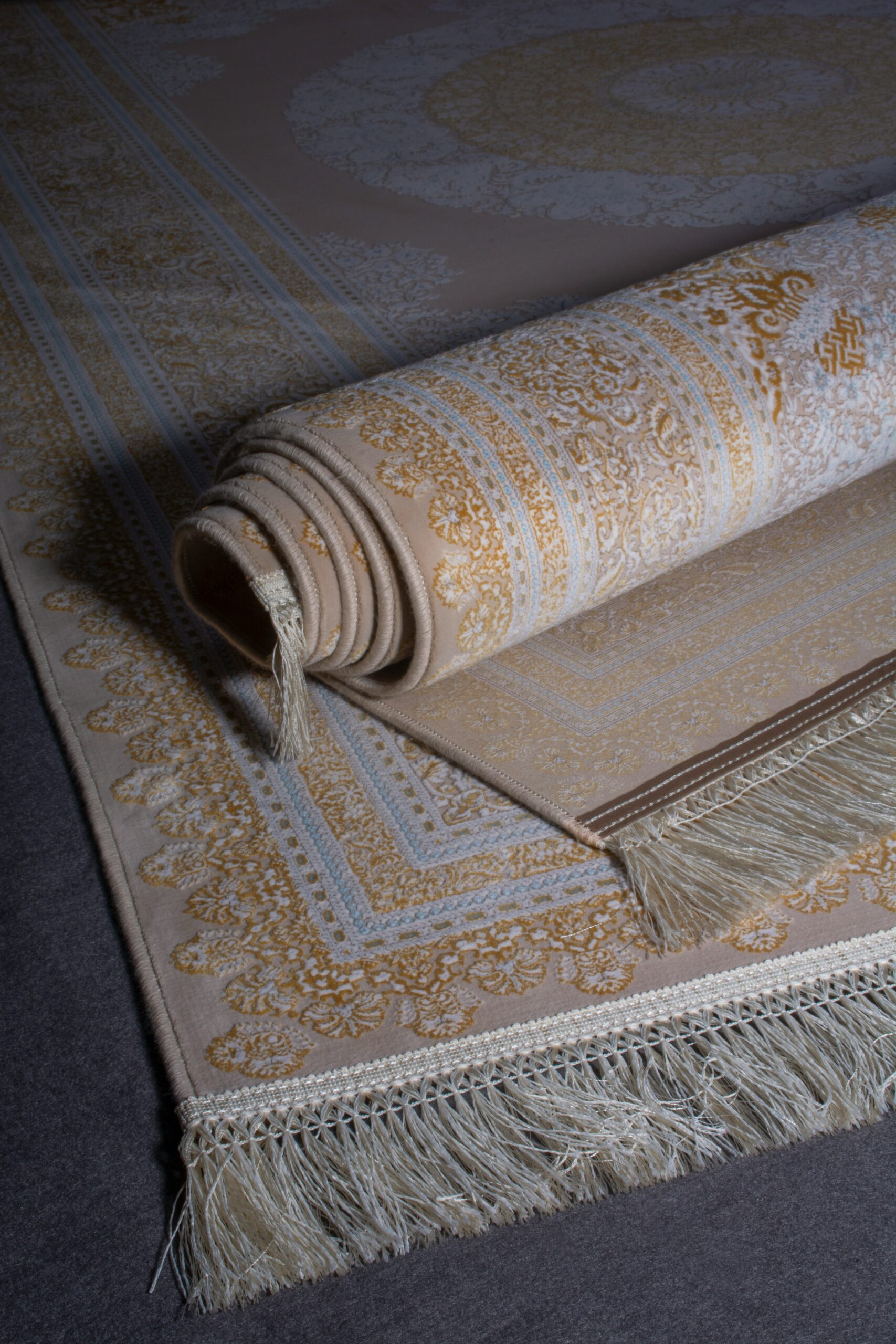 Rolled beige and gold patterned carpet with fringed edges partially unrolled on a dark floor.

