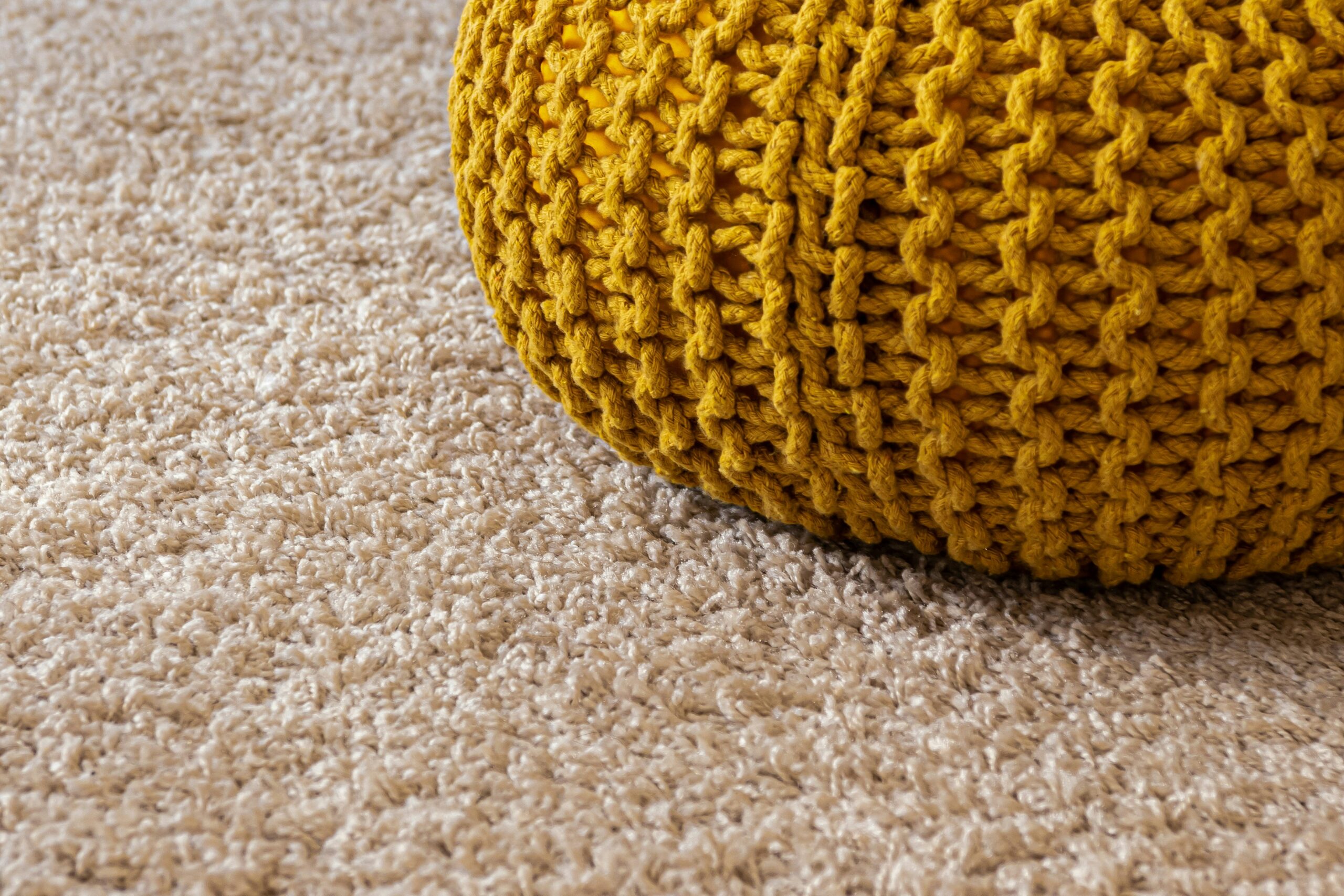 Close-up of beige textured carpet with a mustard yellow knitted pouf in the corner.